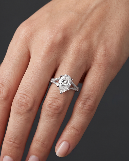 A close-up of a woman's hand wearing a pear-shaped diamond engagement ring with a diamond-encrusted split-shank band. The point of the diamond is facing downward toward her fingertips, set against a dark charcoal gray background.
