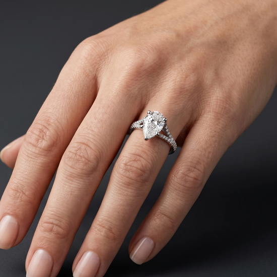 A close-up, editorial-style photograph of a woman's elegantly posed hand wearing a luxury engagement ring. The ring features a large, 3-carat pear-shaped diamond with its point facing the manicured fingernails, set on a white metal split-shank band encrusted with smaller pavé diamonds. The hand is softly lit, showing natural skin texture, against a smooth, dark charcoal gray background that highlights the brilliance and details of the jewelry.