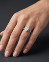 A close-up, editorial-style photograph of a woman's elegantly posed hand wearing a luxury engagement ring. The ring features a large, 3-carat pear-shaped diamond with its point facing the manicured fingernails, set on a white metal split-shank band encrusted with smaller pavé diamonds. The hand is softly lit, showing natural skin texture, against a smooth, dark charcoal gray background that highlights the brilliance and details of the jewelry.