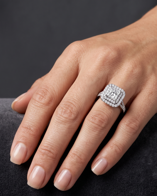 Close-up of a woman's hand resting elegantly on a dark gray surface, wearing a platinum or white gold engagement ring. The ring features a princess-cut center diamond surrounded by a double halo and a diamond-paved split-shank band.