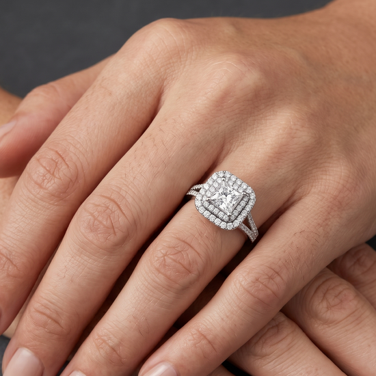 A close-up editorial photo of a woman's hands resting gracefully on a dark charcoal background. On her ring finger, she wears a striking engagement ring featuring a princess-cut center diamond surrounded by a double halo of smaller diamonds.