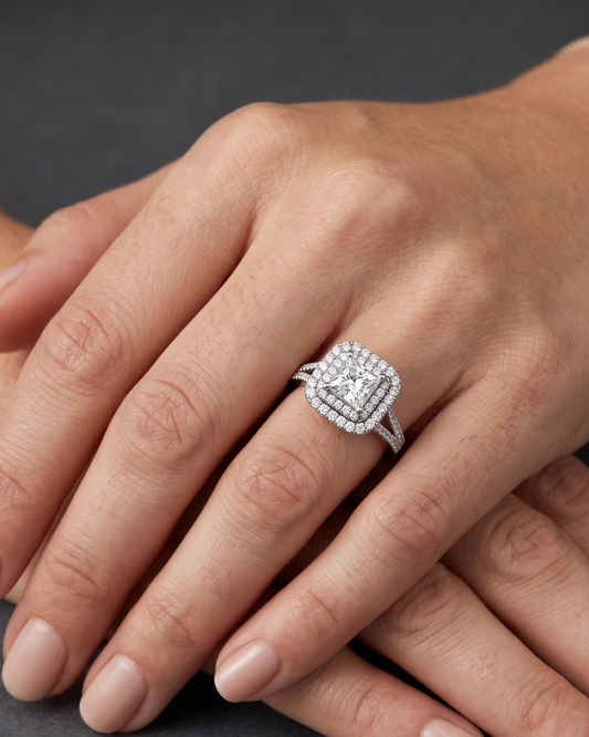 A close-up editorial photo of a woman's hands resting gracefully on a dark charcoal background. On her ring finger, she wears a striking engagement ring featuring a princess-cut center diamond surrounded by a double halo of smaller diamonds.
