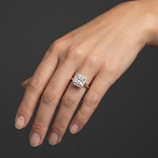 A close-up of a woman's gracefully posed hand wearing a large, princess-cut diamond halo ring against a dark charcoal gray background.