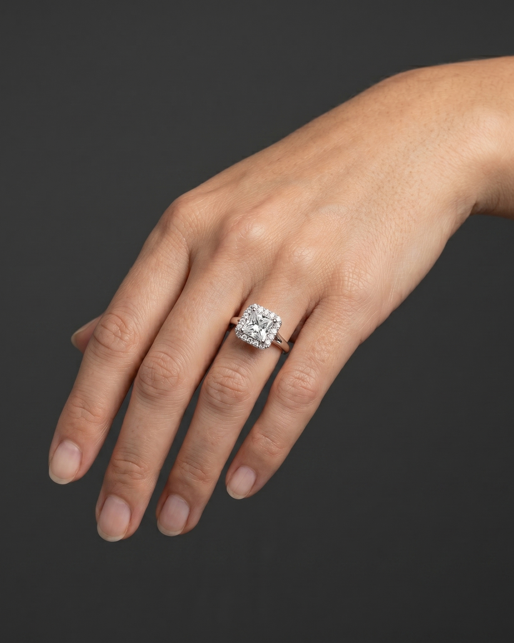 A close-up of a woman's gracefully posed hand wearing a large, princess-cut diamond halo ring against a dark charcoal gray background.