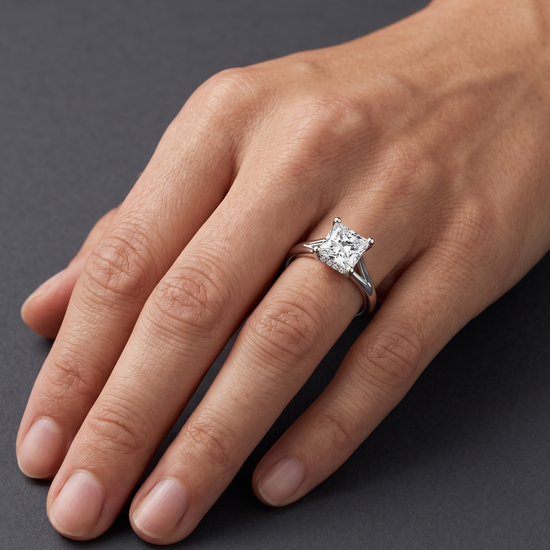 A close-up editorial photo of a woman's hand resting gracefully against a smooth, charcoal gray background. She is wearing a polished white gold or silver engagement ring featuring a large, sparkling princess-cut diamond with a split-shank band.