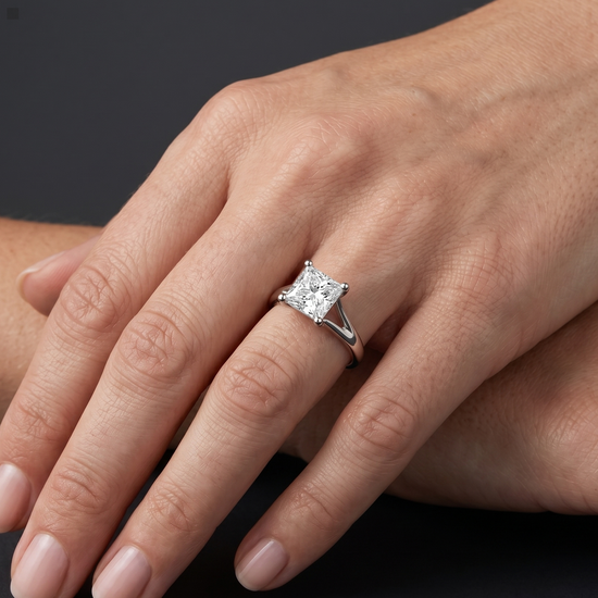 A close-up, hyper-realistic view of a woman's elegantly posed hand wearing a white gold split-shank engagement ring with a prominent princess-cut solitaire diamond. The hand rests softly against the lower arm, displaying natural skin texture and clean manicured nails. Soft, diffused studio lighting highlights the sparkling facets of the diamond and the reflective metal of the band, while a smooth, dark charcoal gray background provides a stark, premium contrast.
