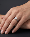 A close-up, hyper-realistic view of a woman's elegantly posed hand wearing a white gold split-shank engagement ring with a prominent princess-cut solitaire diamond. The hand rests softly against the lower arm, displaying natural skin texture and clean manicured nails. Soft, diffused studio lighting highlights the sparkling facets of the diamond and the reflective metal of the band, while a smooth, dark charcoal gray background provides a stark, premium contrast.