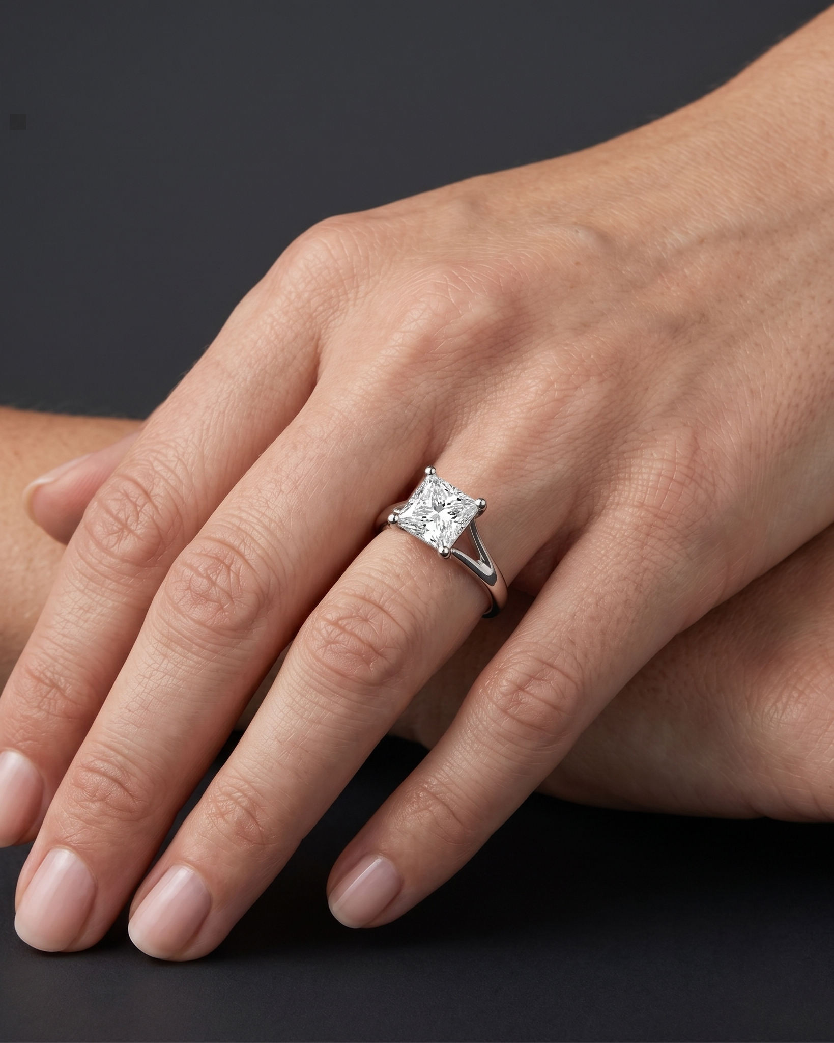 A close-up, hyper-realistic view of a woman's elegantly posed hand wearing a white gold split-shank engagement ring with a prominent princess-cut solitaire diamond. The hand rests softly against the lower arm, displaying natural skin texture and clean manicured nails. Soft, diffused studio lighting highlights the sparkling facets of the diamond and the reflective metal of the band, while a smooth, dark charcoal gray background provides a stark, premium contrast.