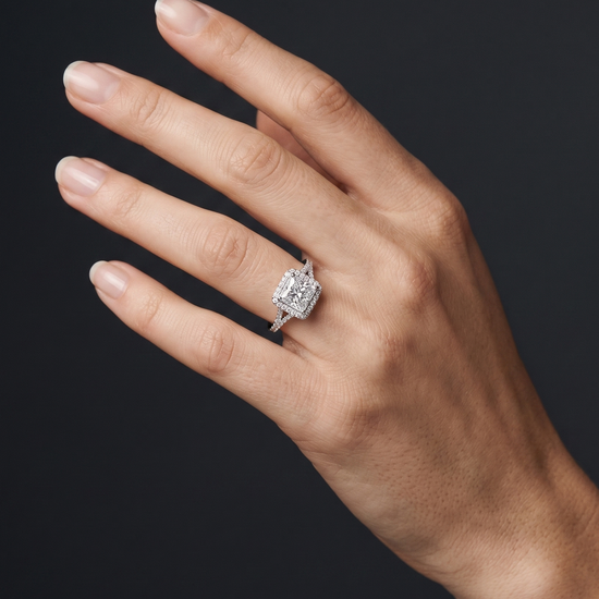 luxury editorial close-up of an elegantly posed woman's hand wearing a silver-toned engagement ring. The ring features a large princess-cut center diamond surrounded by a diamond halo, set on a diamond-encrusted split-shank band. The hand is positioned against a smooth, dark charcoal gray background, with soft studio lighting highlighting the sparkling gemstones and the natural texture of her skin.
