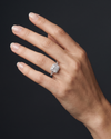luxury editorial close-up of an elegantly posed woman's hand wearing a silver-toned engagement ring. The ring features a large princess-cut center diamond surrounded by a diamond halo, set on a diamond-encrusted split-shank band. The hand is positioned against a smooth, dark charcoal gray background, with soft studio lighting highlighting the sparkling gemstones and the natural texture of her skin.