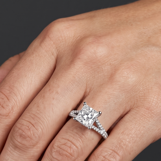 A close-up of a woman's hand wearing a white gold engagement ring against a dark charcoal gray background. The ring features a large princess-cut center diamond and a split-shank band paved with smaller diamonds.