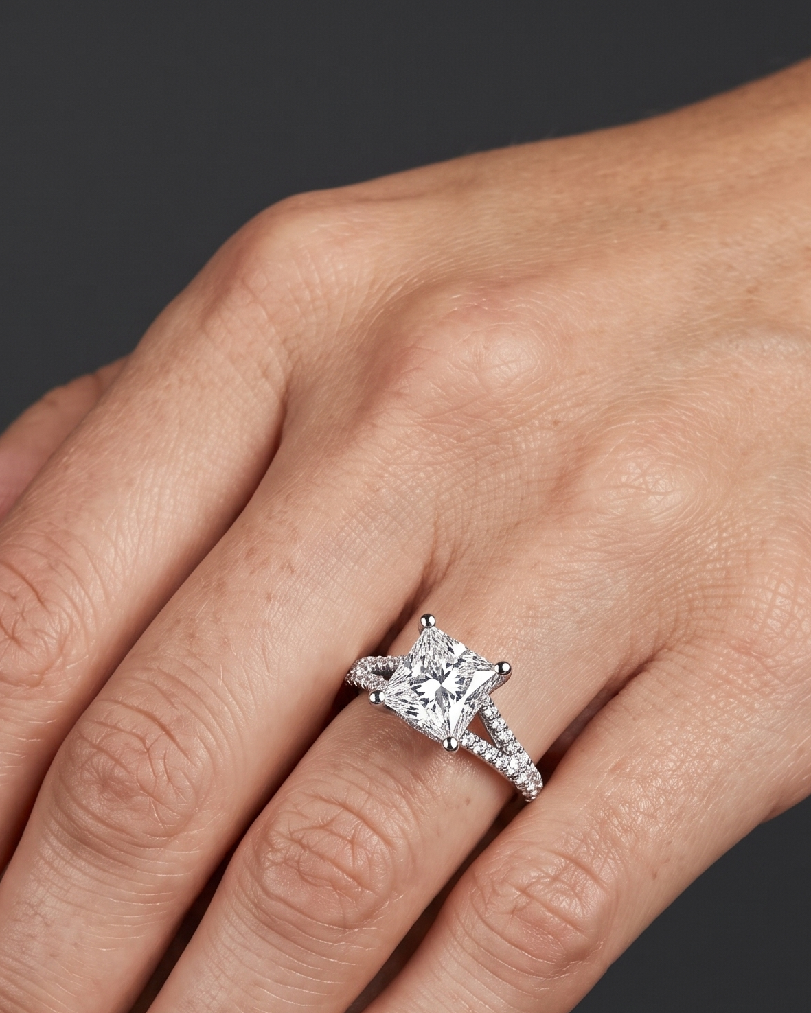 A close-up of a woman's hand wearing a white gold engagement ring against a dark charcoal gray background. The ring features a large princess-cut center diamond and a split-shank band paved with smaller diamonds.