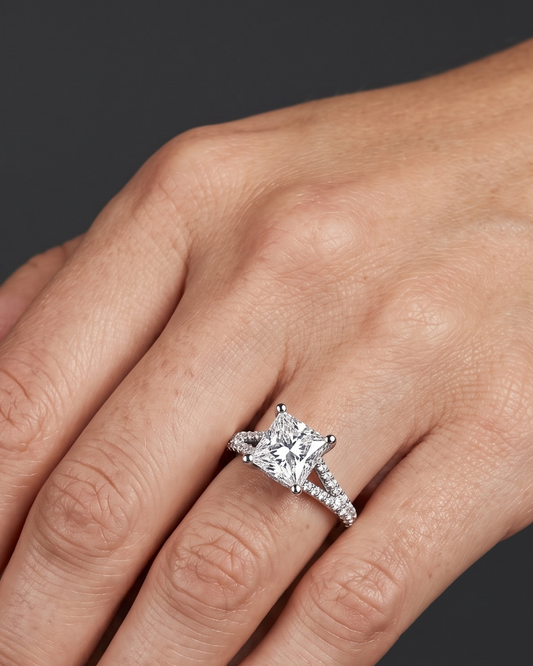 A close-up of a woman's hand wearing a white gold engagement ring against a dark charcoal gray background. The ring features a large princess-cut center diamond and a split-shank band paved with smaller diamonds.