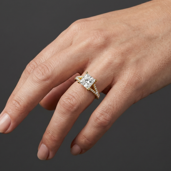 A close-up shot of a gently posed woman's hand resting against a smooth, dark charcoal gray background. On her ring finger, she is wearing a yellow gold engagement ring featuring a large princess-cut center diamond and a diamond-encrusted split shank. Soft studio lighting highlights the sparkle of the gemstones, the polished gold, and the natural texture of her skin.