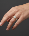 A close-up shot of a gently posed woman's hand resting against a smooth, dark charcoal gray background. On her ring finger, she is wearing a yellow gold engagement ring featuring a large princess-cut center diamond and a diamond-encrusted split shank. Soft studio lighting highlights the sparkle of the gemstones, the polished gold, and the natural texture of her skin.