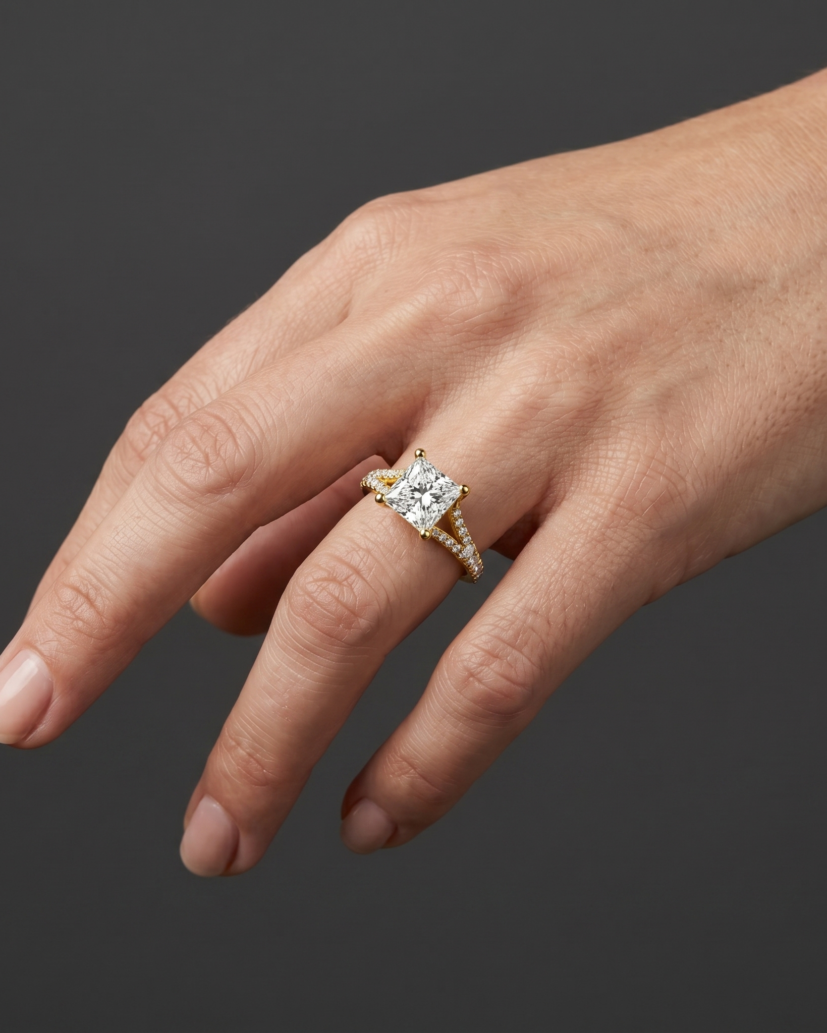 A close-up shot of a gently posed woman's hand resting against a smooth, dark charcoal gray background. On her ring finger, she is wearing a yellow gold engagement ring featuring a large princess-cut center diamond and a diamond-encrusted split shank. Soft studio lighting highlights the sparkle of the gemstones, the polished gold, and the natural texture of her skin.