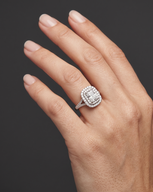 Close-up of a woman's elegantly posed hand wearing a radiant-cut diamond engagement ring with a double halo and split-shank band, set against a dark charcoal gray background.