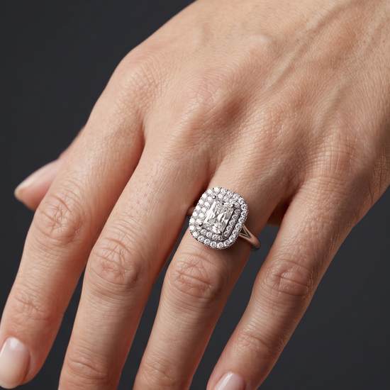 A close-up of a woman's gracefully posed hand against a smooth, dark charcoal gray background, showcasing a luxury engagement ring. The ring features a radiant-cut center diamond surrounded by a double diamond halo on a solid white-metal band.