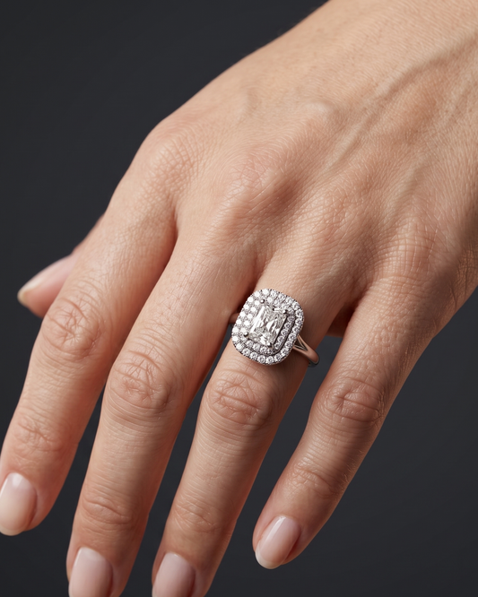 A close-up of a woman's gracefully posed hand against a smooth, dark charcoal gray background, showcasing a luxury engagement ring. The ring features a radiant-cut center diamond surrounded by a double diamond halo on a solid white-metal band.