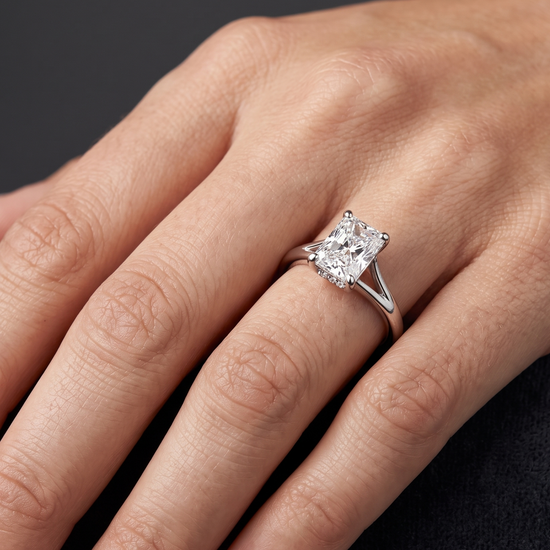 A close-up of a woman's hand elegantly displaying a luxury engagement ring against a smooth, dark charcoal gray background. The ring features a large, radiant-cut center diamond set on a polished silver-toned split-shank band, with a subtle hidden halo of small diamonds visible beneath the main stone. Soft lighting highlights the sparkling facets of the diamond and the natural texture of the hand.