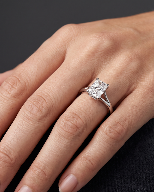 A close-up of a woman's hand elegantly displaying a luxury engagement ring against a smooth, dark charcoal gray background. The ring features a large, radiant-cut center diamond set on a polished silver-toned split-shank band, with a subtle hidden halo of small diamonds visible beneath the main stone. Soft lighting highlights the sparkling facets of the diamond and the natural texture of the hand.