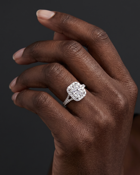 A close-up of a woman's hand with a deep skin tone wearing a sparkling radiant-cut diamond halo engagement ring with a diamond split-shank band, set against a dark gray background.