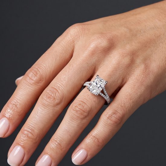 A close-up photograph of a woman's hand wearing a large, sparkling diamond engagement ring on her ring finger. The ring features a rectangular radiant-cut center stone set on a split-shank band encrusted with smaller diamonds. The woman's hand is relaxed and gracefully posed against a smooth, dark charcoal gray background. Her nails are manicured with a pale, natural polish. The lighting highlights the brilliance of the diamonds and the natural texture of her skin.