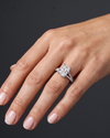 A close-up photograph of a woman's hand wearing a large, sparkling diamond engagement ring on her ring finger. The ring features a rectangular radiant-cut center stone set on a split-shank band encrusted with smaller diamonds. The woman's hand is relaxed and gracefully posed against a smooth, dark charcoal gray background. Her nails are manicured with a pale, natural polish. The lighting highlights the brilliance of the diamonds and the natural texture of her skin.