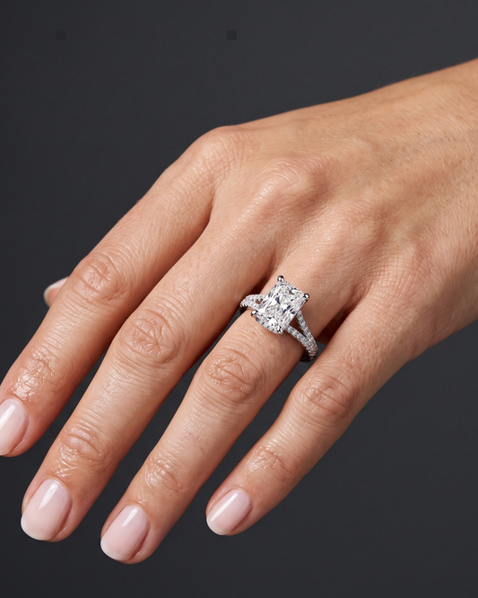 A close-up photograph of a woman's hand wearing a large, sparkling diamond engagement ring on her ring finger. The ring features a rectangular radiant-cut center stone set on a split-shank band encrusted with smaller diamonds. The woman's hand is relaxed and gracefully posed against a smooth, dark charcoal gray background. Her nails are manicured with a pale, natural polish. The lighting highlights the brilliance of the diamonds and the natural texture of her skin.