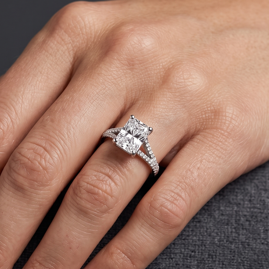 A close-up editorial shot of a woman's hand resting elegantly on a dark charcoal gray textured surface. On her ring finger, she wears a luxury engagement ring featuring a large radiant-cut center diamond and a split-shank band encrusted with smaller pavé diamonds. The lighting is soft and diffused, highlighting the sparkling jewelry, a delicate silver bracelet on her wrist, and the natural texture of her skin.