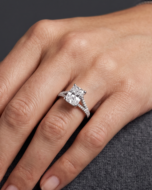 A close-up editorial shot of a woman's hand resting elegantly on a dark charcoal gray textured surface. On her ring finger, she wears a luxury engagement ring featuring a large radiant-cut center diamond and a split-shank band encrusted with smaller pavé diamonds. The lighting is soft and diffused, highlighting the sparkling jewelry, a delicate silver bracelet on her wrist, and the natural texture of her skin.