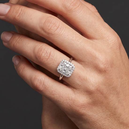 Close-up of a woman's elegantly posed hands against a smooth, dark charcoal gray background. On her ring finger, she is wearing a silver-toned engagement ring featuring a large, radiant-cut center diamond surrounded by a halo of smaller diamonds. Soft lighting highlights the sparkle of the jewelry and the natural texture of her skin.