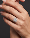 Close-up of a woman's elegantly posed hands against a smooth, dark charcoal gray background. On her ring finger, she is wearing a silver-toned engagement ring featuring a large, radiant-cut center diamond surrounded by a halo of smaller diamonds. Soft lighting highlights the sparkle of the jewelry and the natural texture of her skin.