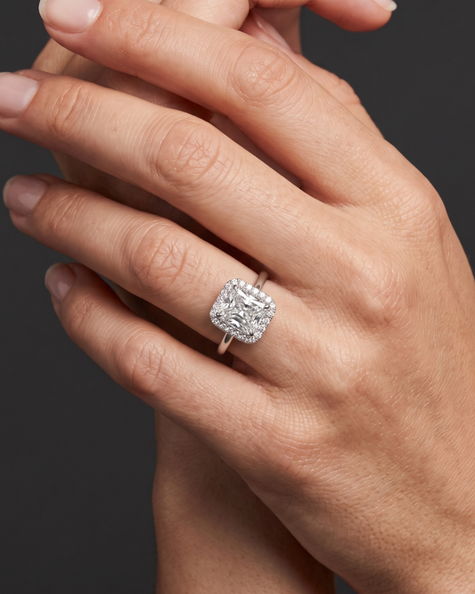Close-up of a woman's elegantly posed hands against a smooth, dark charcoal gray background. On her ring finger, she is wearing a silver-toned engagement ring featuring a large, radiant-cut center diamond surrounded by a halo of smaller diamonds. Soft lighting highlights the sparkle of the jewelry and the natural texture of her skin.