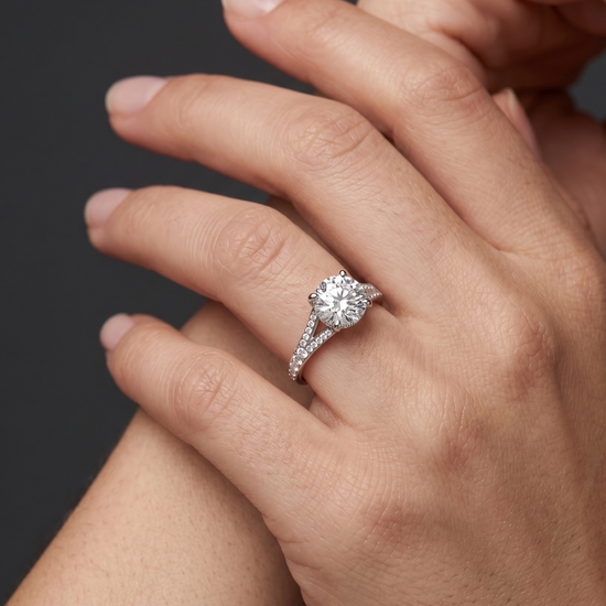 Close-up of delicately posed hands wearing a white gold engagement ring with a large round-cut center diamond and a diamond-encrusted split-shank band, set against a dark gray background.