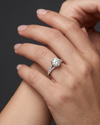 Close-up of delicately posed hands wearing a white gold engagement ring with a large round-cut center diamond and a diamond-encrusted split-shank band, set against a dark gray background.