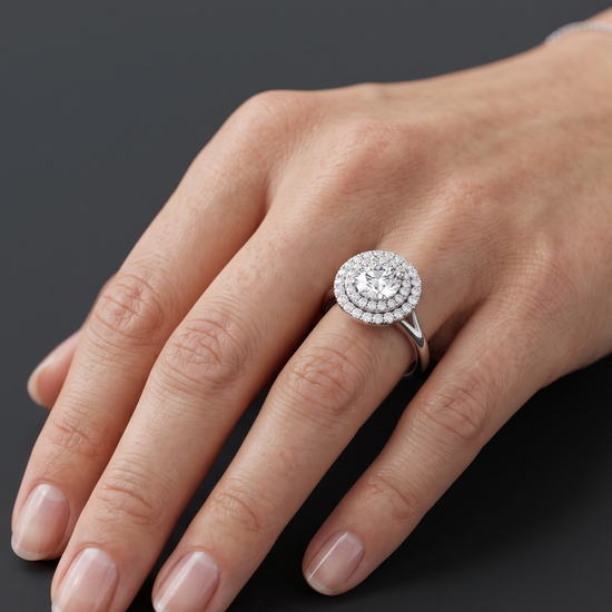 A close-up of a woman's hand resting on a dark charcoal surface, wearing a sparkling silver engagement ring with a large round center diamond and a double halo setting. A delicate silver chain bracelet is visible on her wrist.