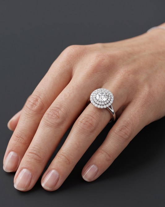 A close-up of a woman's hand resting on a dark charcoal surface, wearing a sparkling silver engagement ring with a large round center diamond and a double halo setting. A delicate silver chain bracelet is visible on her wrist.
