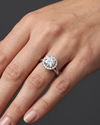 A close-up of a woman's elegantly posed hand wearing a round-cut diamond halo engagement ring on a split-shank band, set against a smooth, dark charcoal gray background.