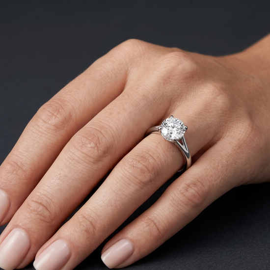 A close-up of a woman's elegantly posed hand wearing a large round-cut diamond engagement ring with a white gold split-shank band, set against a dark charcoal gray background.
