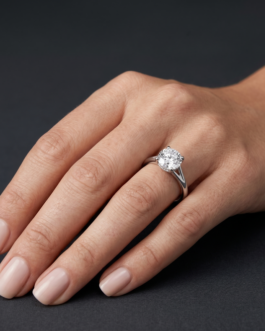 A close-up of a woman's elegantly posed hand wearing a large round-cut diamond engagement ring with a white gold split-shank band, set against a dark charcoal gray background.