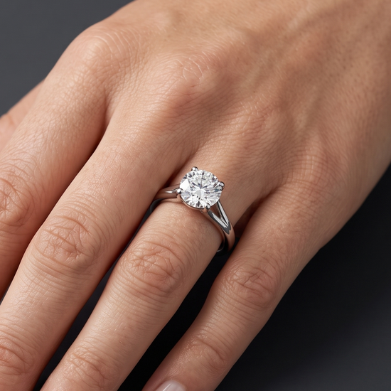 A close-up of a woman's elegantly posed hand resting against a smooth, dark charcoal gray background, wearing a brilliant round-cut diamond solitaire engagement ring on a silver-toned split-shank band.