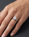 A close-up of a woman's elegantly posed hand resting against a smooth, dark charcoal gray background, wearing a brilliant round-cut diamond solitaire engagement ring on a silver-toned split-shank band.