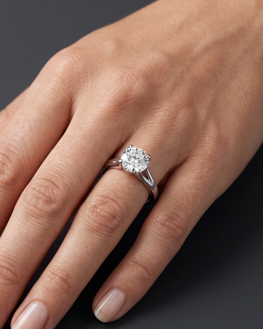 A close-up of a woman's elegantly posed hand resting against a smooth, dark charcoal gray background, wearing a brilliant round-cut diamond solitaire engagement ring on a silver-toned split-shank band.
