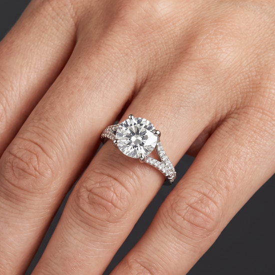 Close-up of a woman's hand resting elegantly against a dark charcoal background, wearing a white gold engagement ring with a round center diamond and a diamond-encrusted split-shank band.