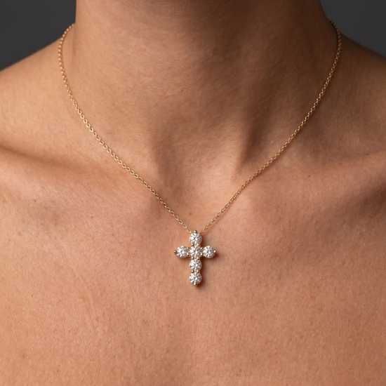 Close-up of a person wearing a delicate gold chain necklace with a cross pendant made of six sparkling round diamonds, resting on their bare chest against a dark background.