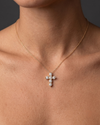 Close-up of a person wearing a delicate gold chain necklace with a cross pendant made of six sparkling round diamonds, resting on their bare chest against a dark background.