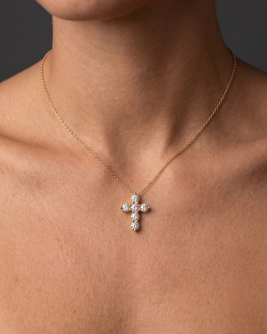 Close-up of a person wearing a delicate gold chain necklace with a cross pendant made of six sparkling round diamonds, resting on their bare chest against a dark background.