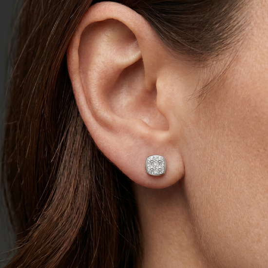 A close-up, editorial-style photograph focusing on a woman's ear and jawline to showcase a petite, cushion-shaped stud earring. The silver-toned earring features a cluster of sparkling clear gemstones set within a smooth metallic border. The model has dark brown hair tucked behind her ear, set against a seamless dark charcoal gray background.