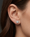 A close-up, editorial-style photograph focusing on a woman's ear and jawline to showcase a petite, cushion-shaped stud earring. The silver-toned earring features a cluster of sparkling clear gemstones set within a smooth metallic border. The model has dark brown hair tucked behind her ear, set against a seamless dark charcoal gray background.